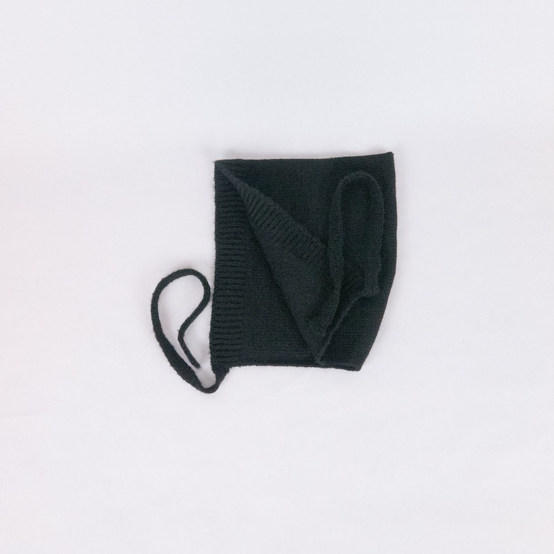 May include: Black knitted baby bonnet with a tie string. The bonnet is folded in half and the tie string is looped around the top.
