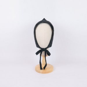 May include: A black knitted baby bonnet with a tie. The bonnet is on a wooden display stand.