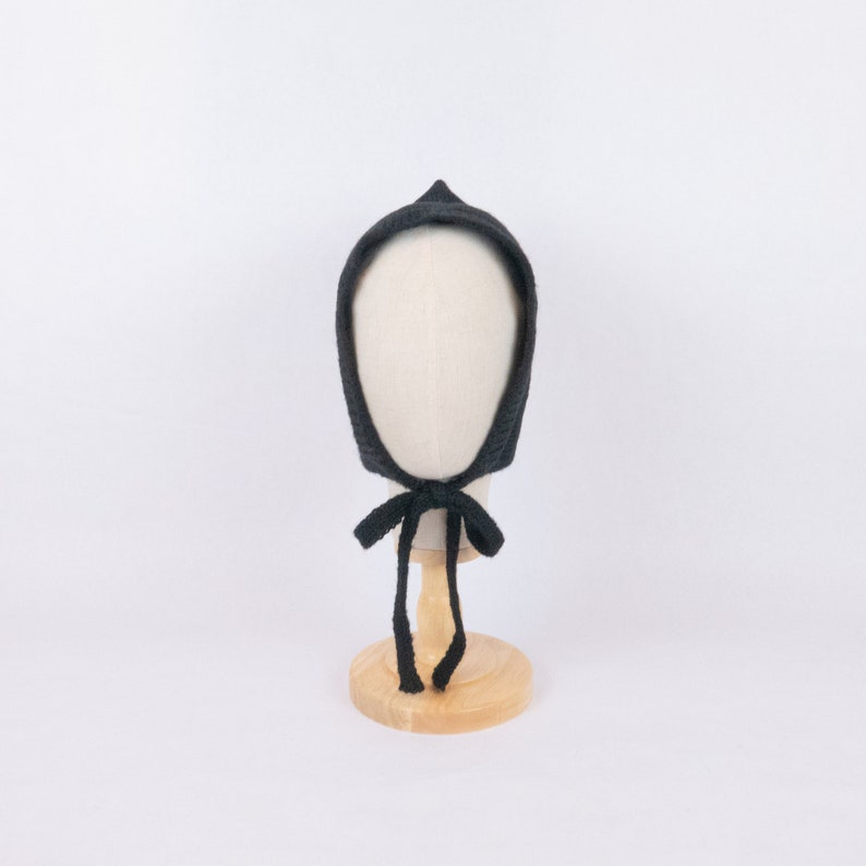 May include: A black knitted baby bonnet with a tie. The bonnet is on a wooden display stand.