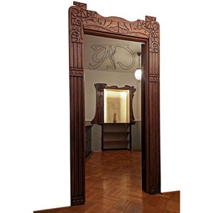 May include: A dark wood doorway with an ornate carved frame. The frame features intricate designs and a decorative top. The doorway leads into a room with a wooden floor and a shelf unit.