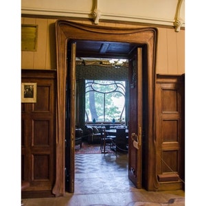 May include: A wooden doorway with an ornate, curved frame leads into a room with a large window and a patterned floor. The room is furnished with a sofa, chairs, and a table.