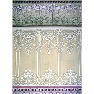 May include: A close-up of a wall with a decorative pattern of white flowers and vines on a light beige background. The pattern is repeated along the wall, creating a symmetrical design. The bottom of the wall features a row of pink and green tiles.