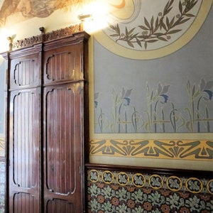 Puede incluir: Un par de puertas dobles de madera oscura con tallas intrincadas, colocadas contra una pared con un mural decorativo de estilo art nouveau que presenta motivos florales y un fondo azul pálido.