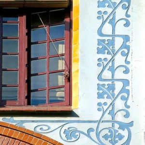 May include: A brick building with a window and a decorative blue and white floral design on the wall. The window has a brown wooden frame and a brown blind.