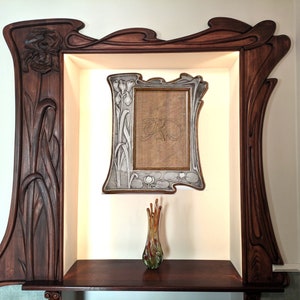 May include: A dark wood frame with intricate carvings of flowers and leaves surrounds a smaller, silver-toned frame with a drawing inside. A vase with a green and brown design sits on a dark wood shelf below the frames.