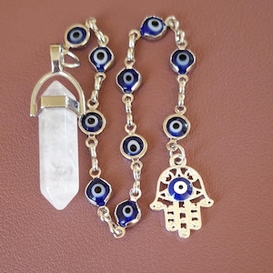 May include: A silver necklace featuring blue evil eye beads and a Hamsa hand charm. A clear quartz crystal pendant is on the left. The necklace is displayed on a brown surface.