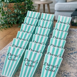 May include: A collection of popcorn boxes, each featuring a teal and white striped pattern. The boxes are inscribed with "Darryl's Class" in black script. The arrangement suggests a party or event, with the boxes neatly aligned.