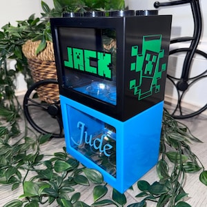 Peut inclure: Une veilleuse personnalisée à deux niveaux avec les noms "Jack" et "Jude" affichés. La partie supérieure est noire avec du texte vert et un personnage pixélisé, tandis que le bas est bleu. Le design ressemble à une brique de construction.