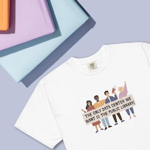 May include: A white short-sleeved t-shirt featuring a graphic illustration of four diverse individuals holding a banner that reads, THE ONLY DATA CENTER WE WANT IS THE PUBLIC LIBRARY! The shirt has a crew neckline and a visible interior brand label.