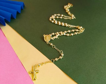 May include: A gold-toned rosary necklace with a small, round pendant featuring a religious image. The necklace is on a multi-colored background.