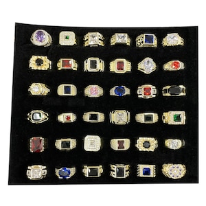 May include: A collection of gold-toned rings with various gemstone settings, including emerald, ruby, sapphire, and diamond-like stones. The rings are displayed on a black velvet surface, showcasing their intricate designs and vibrant colors.