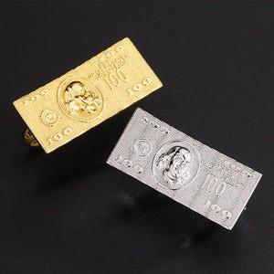 May include: Two rectangular cufflinks, one gold-toned and one silver-toned, each resembling a U.S. $100 bill. The cufflinks feature a portrait and the number 100. The background is a dark, solid color.