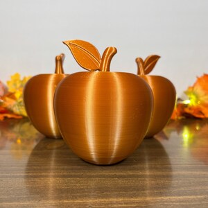 May include: Three bronze-colored apple-shaped decorative objects with stems and leaves. The apples are arranged in a row, with the central one in focus. The background features fall foliage and soft lighting, creating a seasonal theme.