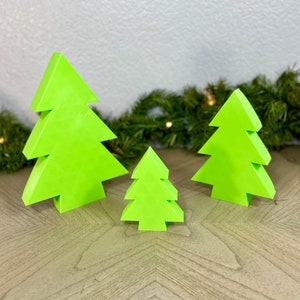 May include: Three neon green wooden Christmas tree decorations of varying sizes. The trees are arranged in a row on a wooden surface, with a garland of greenery in the background. A small silver tree is also visible.