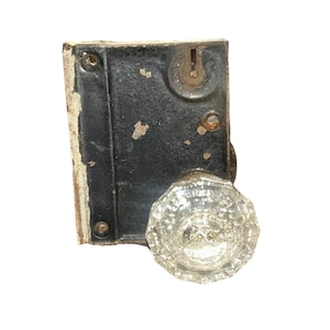 May include: A vintage door lock mechanism featuring a rectangular black metal plate with visible paint wear and a keyhole. Attached to the plate is a clear, faceted glass doorknob with an octagonal shape and a metallic center mounting point.
