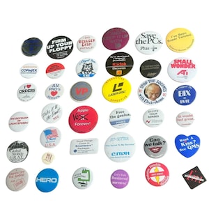 May include: A collection of various round and shaped pin-back buttons featuring diverse colors, graphics, and text including phrases like Firm Up Your Floppy, Save the PCs, Apple 16 Forever, and Laser Disc, displayed in a scattered arrangement.