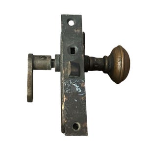 May include: A weathered metal mortise lock mechanism featuring a dark, oxidized finish. The hardware includes a rounded brass knob on one side, a flat lever handle on the opposite side, and a rectangular faceplate with two screw holes and a latch bolt.