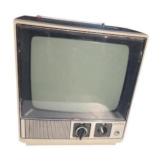 May include: A vintage Sony portable television with a beige plastic casing and a dark screen. The front panel features a speaker grille, two black rotary control knobs, and a small power indicator light. A metal telescoping antenna is attached to the top.