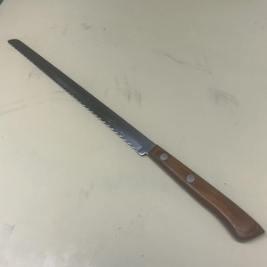 May include: A long, serrated bread knife with a dark metal blade and a wooden handle. The handle is secured with two silver rivets. The knife is designed for slicing bread and other baked goods.