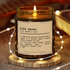 Puede incluir: Una vela encendida en un tarro de vidrio con la etiqueta "Life Saver". La vela está sobre un posavasos de madera, rodeada de luces de cadena. El tarro está lleno de cera oscura.
