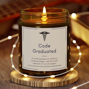 May include: A brown glass candle with a white label that reads "Code Graduated". The label also features a medical symbol and the text "A soothing blend of caffeine, textbook pages, and the sweet absence of on-call pagers".