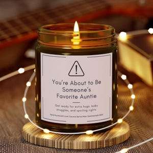 May include: A lit candle in a glass jar with a gold lid. The label reads "You're About to Be Someone's Favorite Auntie." The candle sits on a wooden coaster, surrounded by string lights. The text below the title says "Get ready for extra hugs, baby snuggles, and spoiling rights."