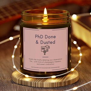 May include: A brown glass candle jar with a pink label that reads "PhD Done & Dusted". The label also includes a small illustration of a graduation cap and diploma. The candle is lit and the text on the label reads "Smells like finally deleting your dissertation file... with hints of champagne, freedom, and sweet vindication."