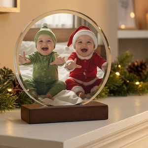 May include: A clear, circular photo frame with two babies dressed in Christmas outfits. One baby is in a green outfit and hat, the other in a red Santa suit and hat. The frame sits on a wooden base.