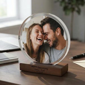 May include: A clear acrylic photo display with a circular frame, featuring a smiling couple. The display is set on a wooden base engraved with "HAPPY Anniversary" and names. A laptop, pen, and envelope are in the background.
