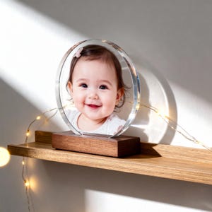 May include: A clear, circular photo frame with a smiling baby's photo inside, set on a wooden base. The frame is illuminated by a string of small, warm-toned lights. The frame sits on a wooden shelf.