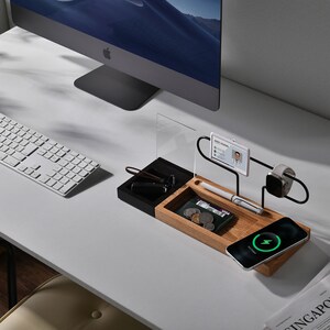 May include: A wooden and black desktop organiser with a charging pad for a phone, a watch stand, a pen holder, and a compartment for small items. The organiser is on a white desk with a keyboard and a computer monitor.