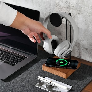 May include: A white wireless headset with a black charging pad and a laptop in the background. The charging pad has a green lightning bolt icon on the screen.