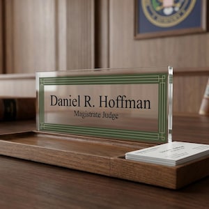 May include: A wooden desk nameplate with the name "Daniel R. Hoffman" and title "Magistrate Judge" displayed on a clear acrylic panel with a green border. A wooden tray holds business cards. The background includes books and a framed emblem.