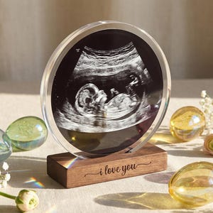 May include: A clear acrylic ultrasound photo display with a wooden base. The ultrasound image shows a baby. The base is engraved with the words "i love you". Decorative glass eggs and flowers surround the display.