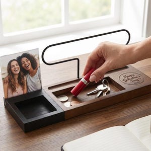 May include: A wooden desk organizer with a black and brown finish. It features a photo frame with two women, a small tray for coins, and a section for keys. A red lipstick is being placed in the tray. The words "Happy Galentine's Day" are engraved on the wood.