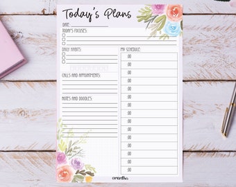 May include: A white planner page with the text "Today's Plans" and floral watercolor accents in pink, orange, and blue. The page includes sections for date, focus, habits, schedule, calls, and notes. A pink notebook and a silver pen are also visible.
