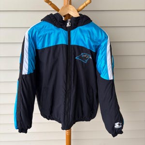 May include: A black, blue, and white hooded jacket with a full zipper. The jacket features the Carolina Panthers logo on the chest and white stripes down the sleeves. The Starter logo is visible on the sleeve.