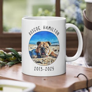 May include: White ceramic mug featuring a color photo of a bulldog on a beach, with the name "Roscoe Hamilton" and the years "2013-2025" printed on it. The mug has a curved handle and is sitting on a wooden surface.
