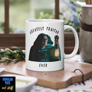 Könnte beinhalten: Weiße Keramiktasse mit dem Aufdruck "GREATEST TRAITOR EVER". Die Tasse zeigt das Bild einer Person in einem Kapuzenmantel, die eine Laterne hält. Die Tasse hat einen großen Henkel und steht auf einer Holzoberfläche.
