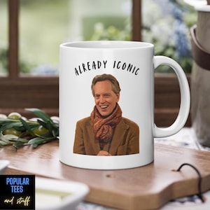 May include: A white ceramic mug featuring a printed image of a smiling person wearing a brown blazer and a patterned orange scarf. Above the person, the text ALREADY ICONIC is printed in a black, handwritten-style font. The mug has a handle.
