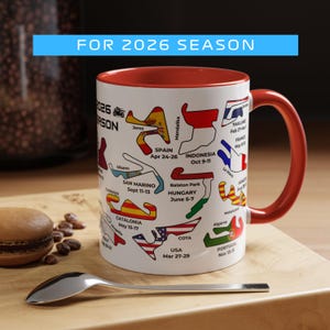 May include: A white ceramic mug with a red interior and handle. The mug features colorful race track designs and text indicating the 2026 season, including locations like Spain and the USA. A spoon and a pastry are on a wooden surface.