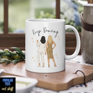 Może przedstawiać: Biały ceramiczny kubek z napisem "Keep Dancing" i ilustracją dwóch kobiet. Jedna ma na sobie biały garnitur, druga złotą sukienkę. Kubek ma duży uchwyt i stoi na drewnianej powierzchni.