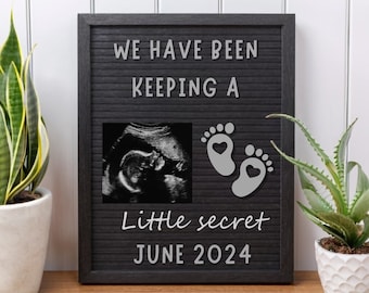 Puede incluir: Un tablero de letras negro con letras blancas que deletrean "We have been keeping a little secret June 2024". Una imagen de ultrasonido en blanco y negro y dos huellas de pies grises con corazones se muestran en el tablero.