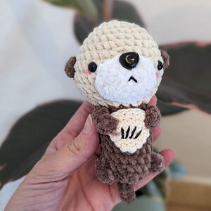 Peut inclure: Un jouet en peluche de loutre de mer en crochet avec une fourrure brune et un ventre blanc. La loutre tient une petite coquille en crochet dans ses pattes. Le jouet a des yeux noirs et un nez rose.