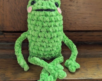 Op de afbeelding: Een groene gehaakte kikker met zwarte ogen en roze wangen. De kikker zit op een houten oppervlak.