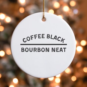 May include: A white, round ceramic ornament with the words "COFFEE BLACK" above a horizontal line, and "BOURBON NEAT" below. The ornament hangs from a gold-colored string, with a blurred background of warm, out-of-focus lights.