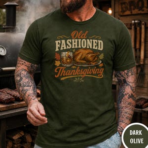 Op de afbeelding: Een donker olijfgroen t-shirt met een grafische print met de tekst Old Fashioned Thanksgiving. Het ontwerp bevat een illustratie van een gebraden kalkoen op een schaal naast een glas whisky, omringd door herfstbladeren in oranje en geel.