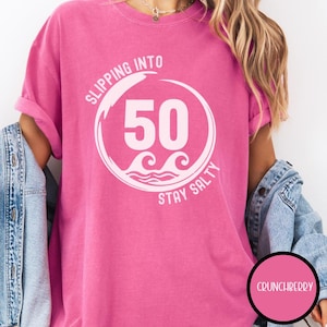 May include: A pink t-shirt with the text "SLIPPING INTO 50 STAY SALTY" in white. The number 50 is in a white circle with waves at the bottom. The shirt is a solid pink color.