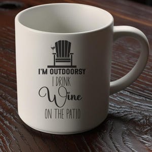 May include: White ceramic mug with a black graphic of an Adirondack chair and wine glass. The mug has the text "I'M OUTDOORSY I DRINK Wine ON THE PATIO" in black script and block letters. The mug has a curved handle.
