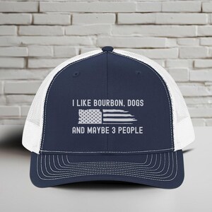 Puede incluir: Gorra de camionero azul marino y blanca con el texto "I LIKE BOURBON, DOGS AND MAYBE 3 PEOPLE" y una bandera americana desgastada. La gorra tiene una parte trasera de malla y una visera curva.